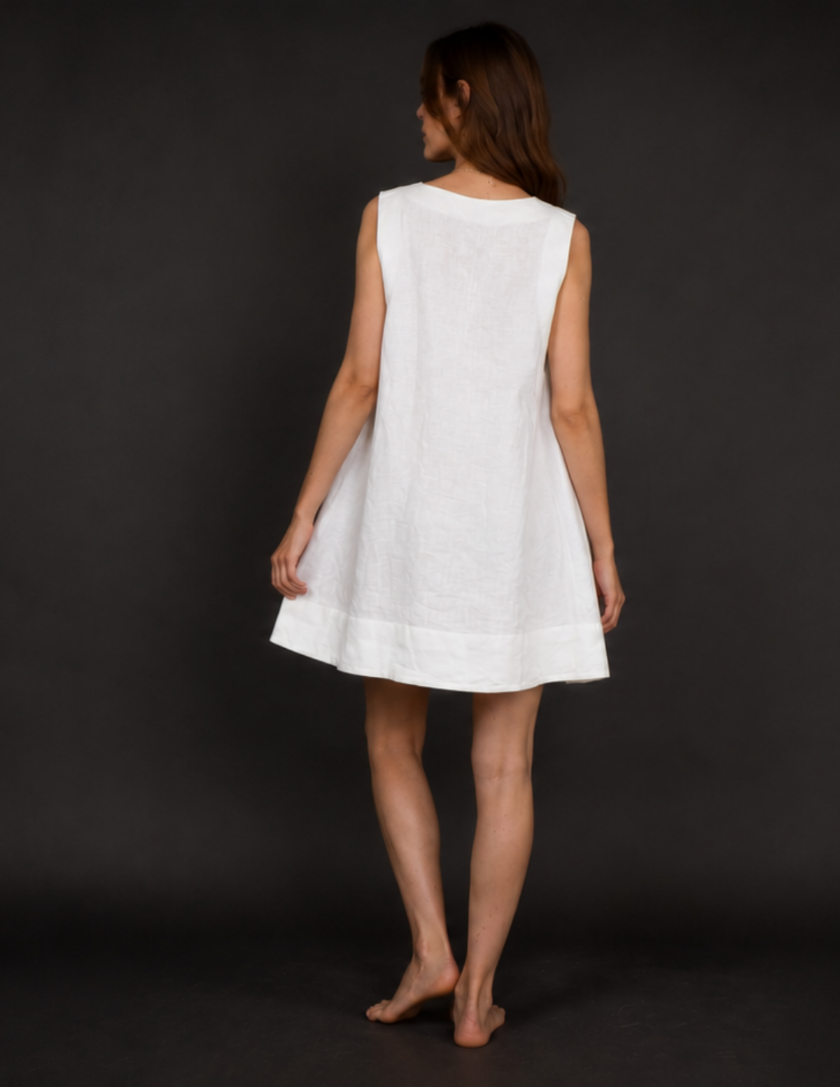 A woman wearing a sleeveless white linen dress, shown from the back against a dark gray studio background.