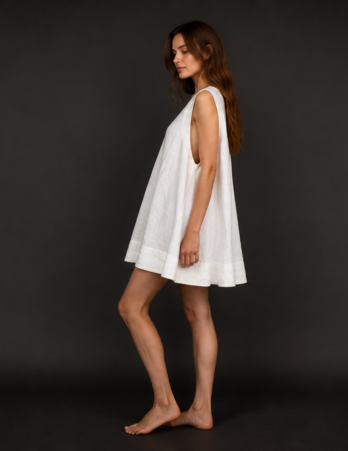 A woman modeling a white sleeveless linen nightgown or tunic dress, shown in profile against a dark gray studio background.