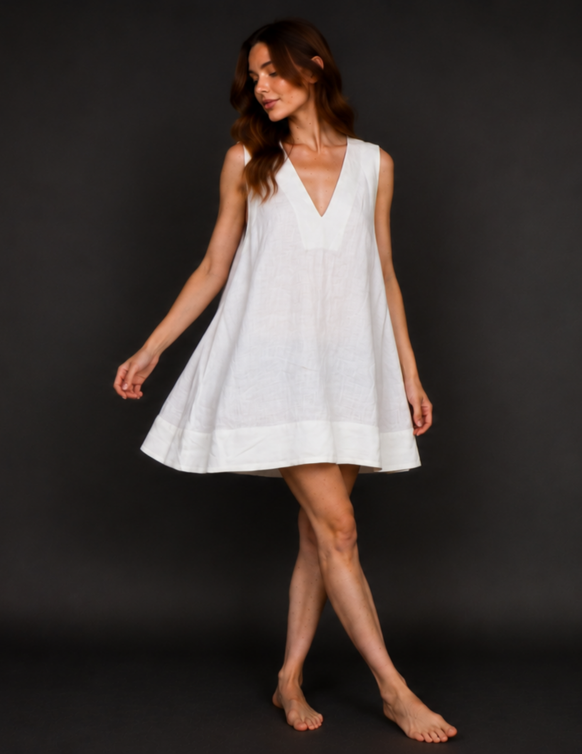 A woman modeling a sleeveless white linen tunic dress with a V-neckline, photographed against a dark gray studio background.