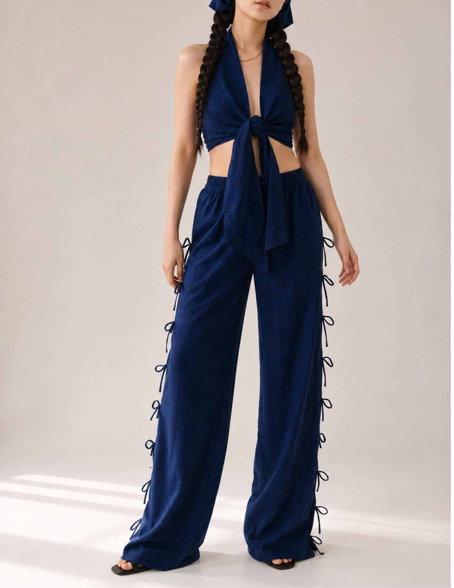 A model wearing a navy blue plaid halter crop top and matching wide-leg pants with side laces, styled with a matching headband and black sandals against a neutral studio background.