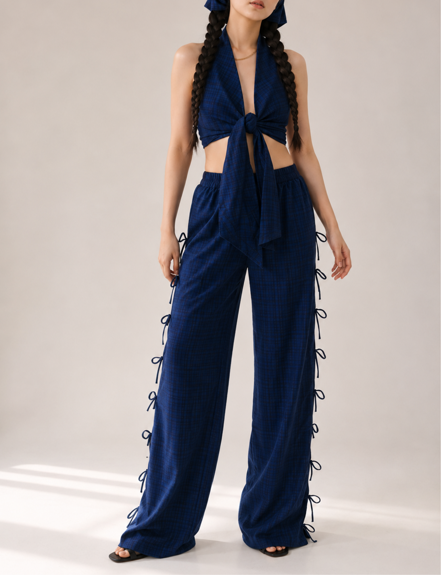 A model wearing a navy blue plaid halter crop top and matching wide-leg pants with side laces, styled with a matching headband and black sandals against a neutral studio background.