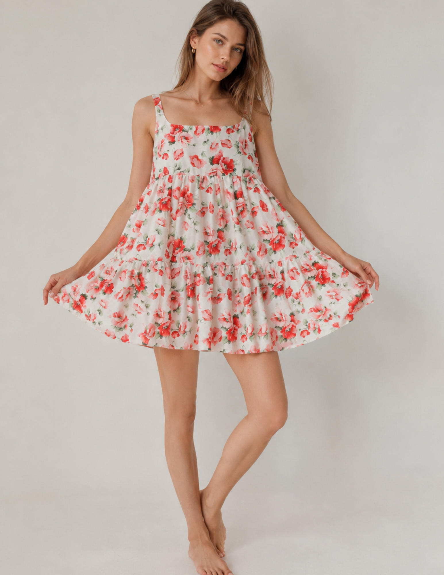 A woman modeling a sleeveless white tiered mini dress with a red and green floral print, standing barefoot against a light gray studio background.