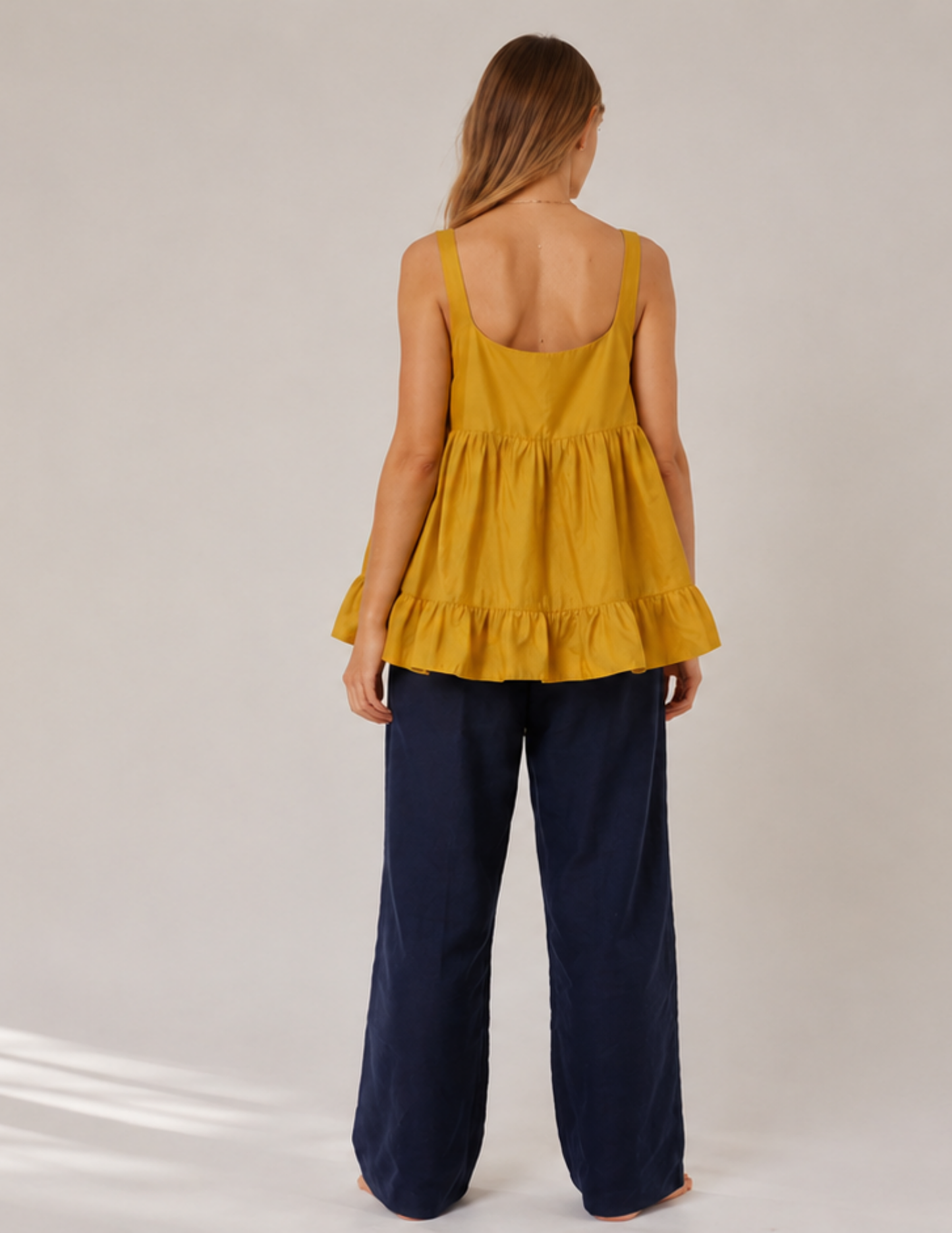 A woman shown from the back wearing a mustard yellow sleeveless peplum top and dark navy wide-leg pants, standing against a light gray studio background.