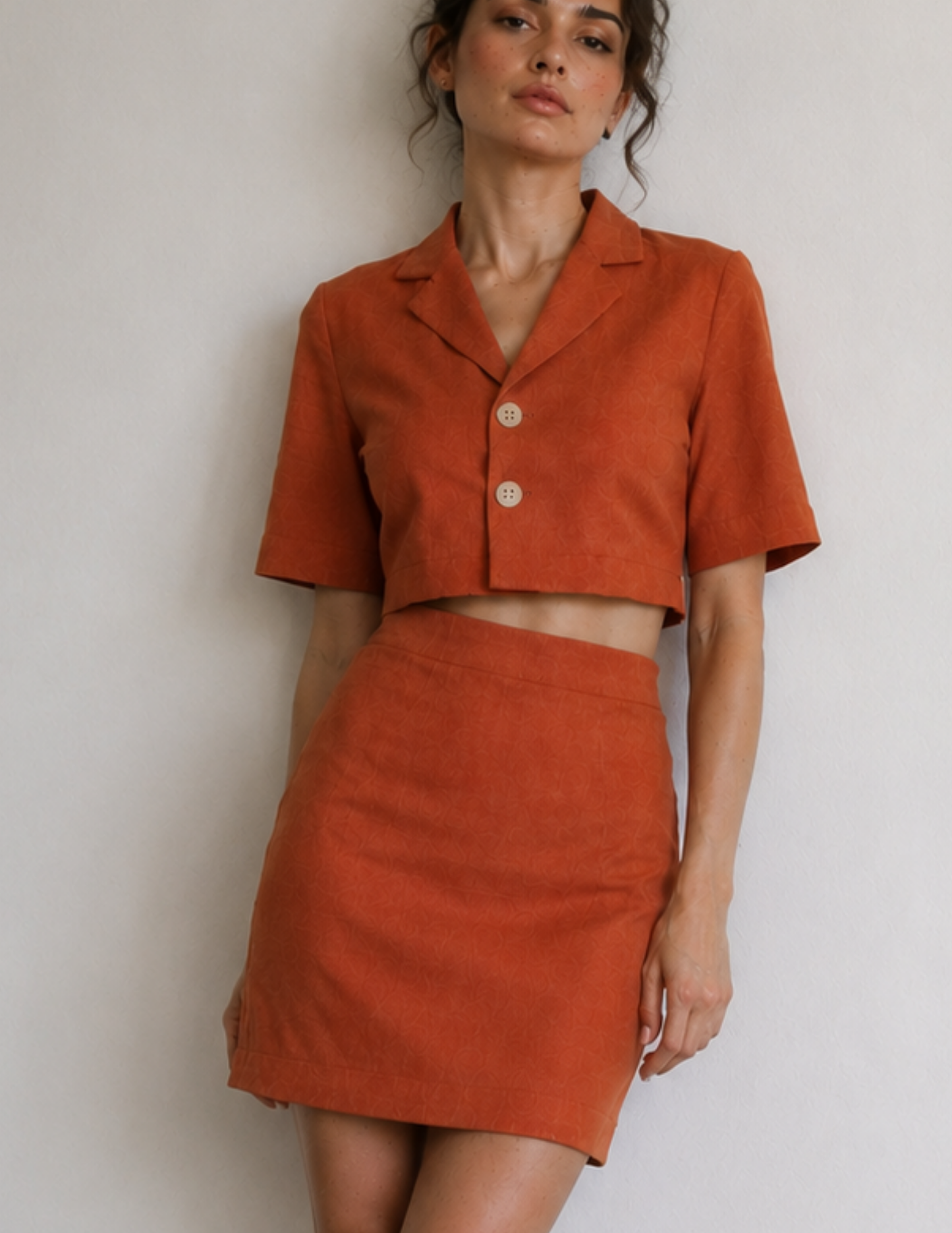 A woman modeling a matching rust-orange two-piece set consisting of a short-sleeve cropped button-front top and a high-waisted mini skirt, set against a plain light gray wall.