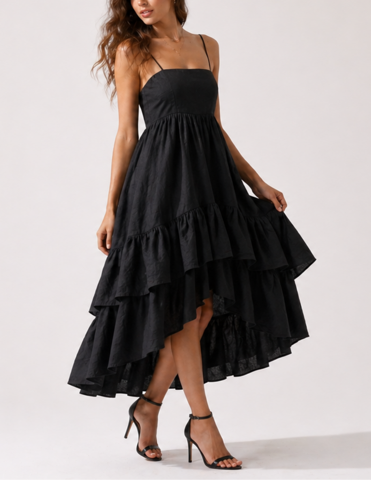 A woman modeling a black high-low tiered midi dress with spaghetti straps and black strappy high-heeled sandals against a plain light gray studio background.