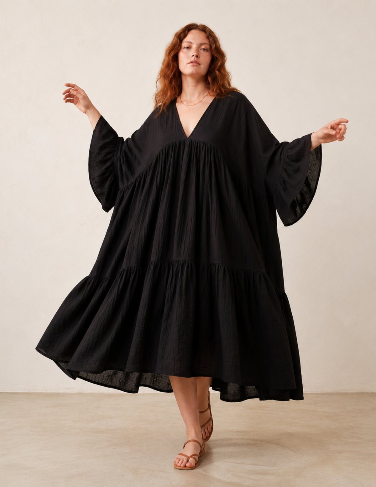 A woman modeling a loose, tiered black dress with wide bell sleeves and a deep V-neckline, photographed against a plain beige studio backdrop.