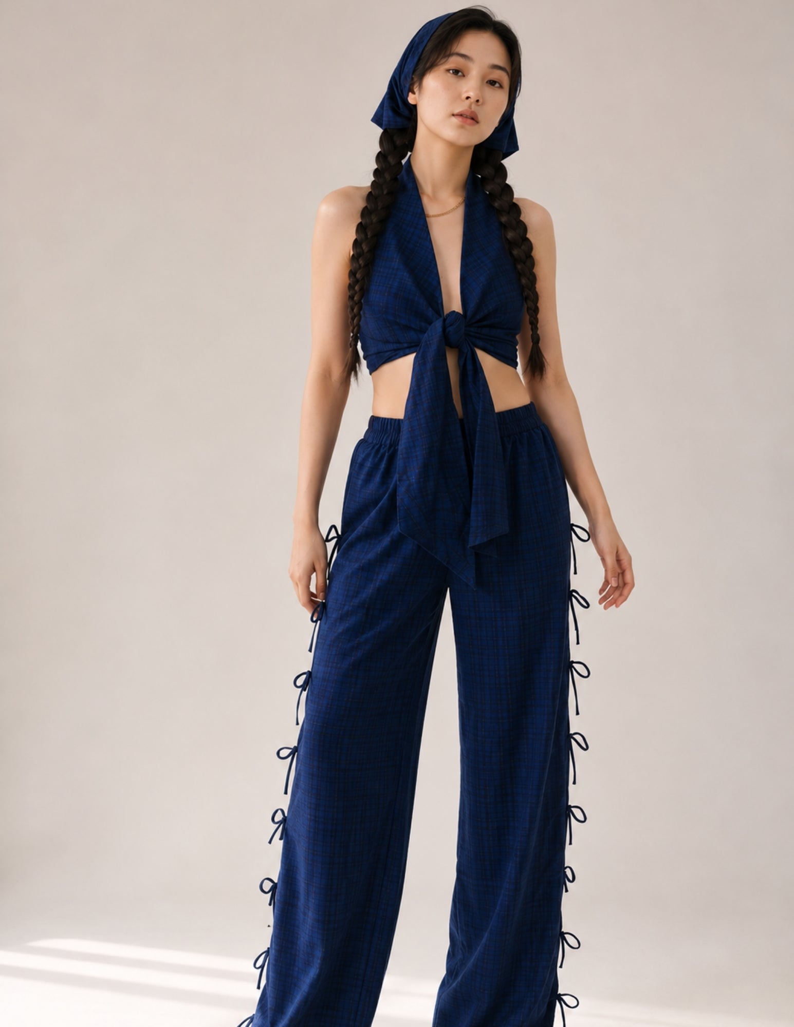 A model wearing a dark blue plaid two-piece outfit consisting of a halter crop top with a front knot and matching wide-leg pants with side laces, along with a matching headscarf, set against a plain light beige studio background.
