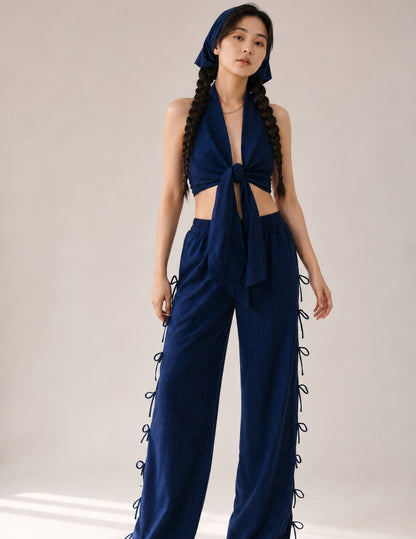 A model wearing a dark blue plaid two-piece outfit consisting of a halter crop top with a front knot and matching wide-leg pants with side laces, along with a matching headscarf, set against a plain light beige studio background.