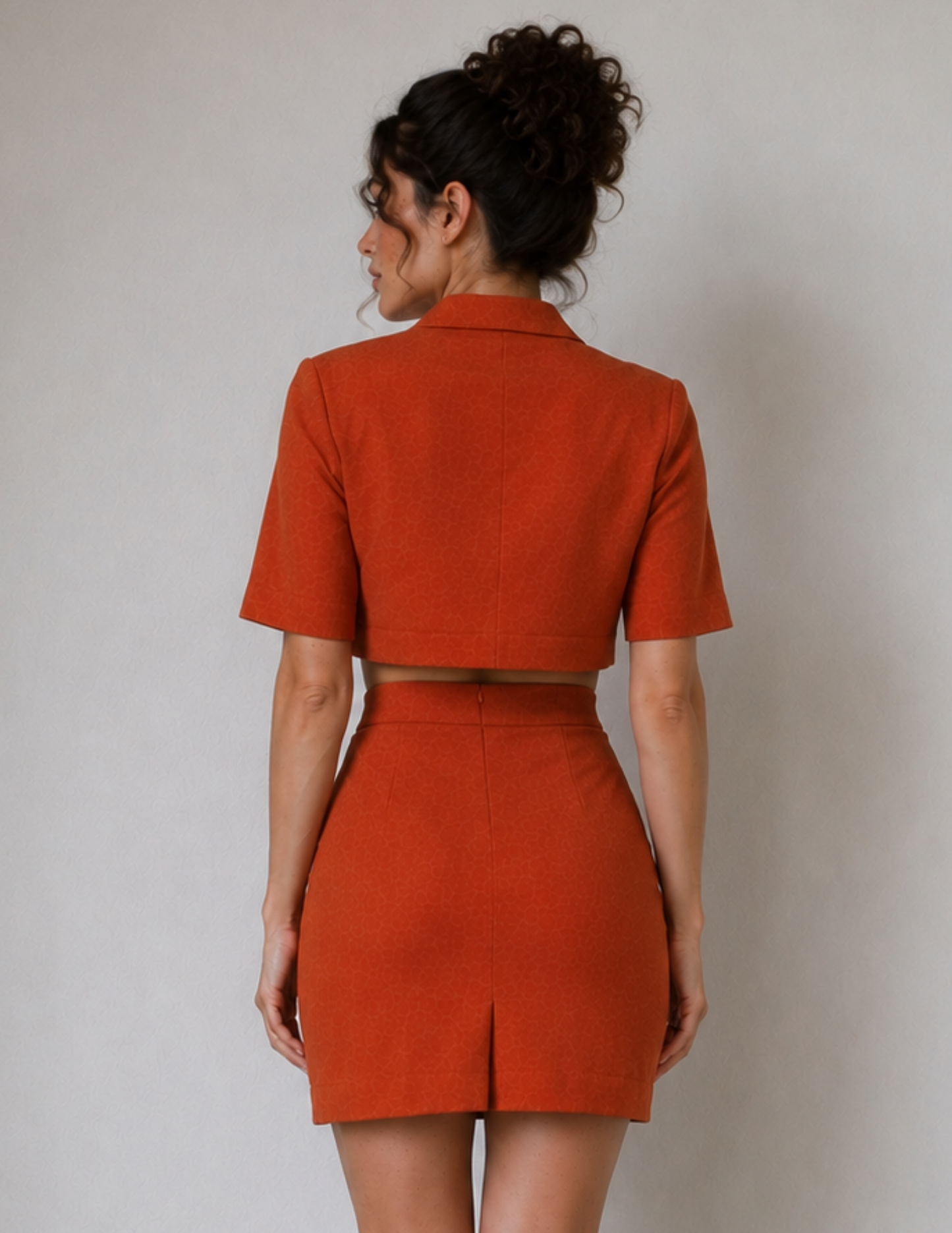 A woman shown from the back wearing a matching rust-orange cropped blazer and high-waisted mini skirt set against a plain light gray studio background.