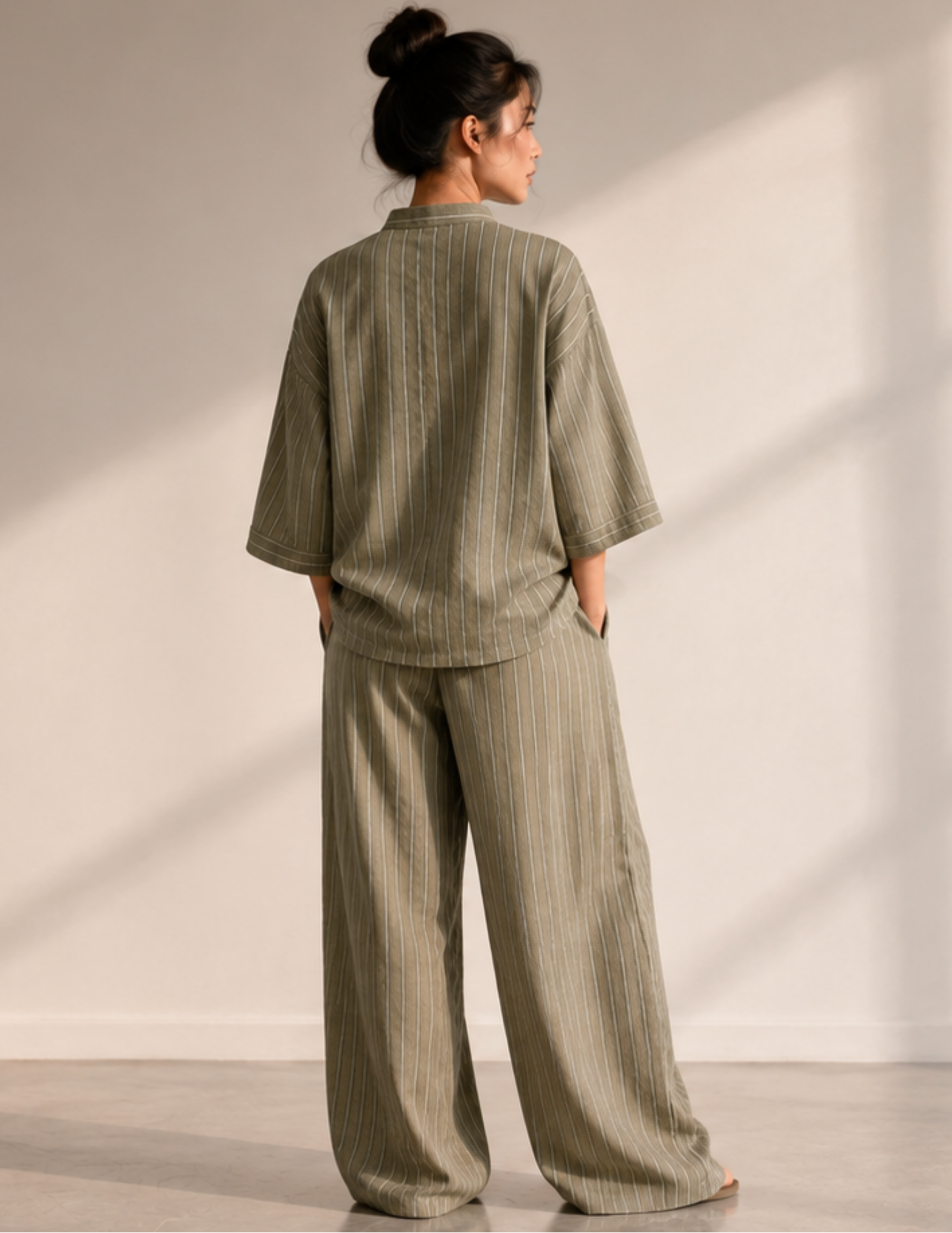 A woman stands with her back to the camera wearing a matching two-piece lounge set in a muted sage green color with thin vertical stripes. The set includes a short-sleeve top and wide-leg pants, photographed in a minimalist room with warm natural light casting soft shadows on the wall.