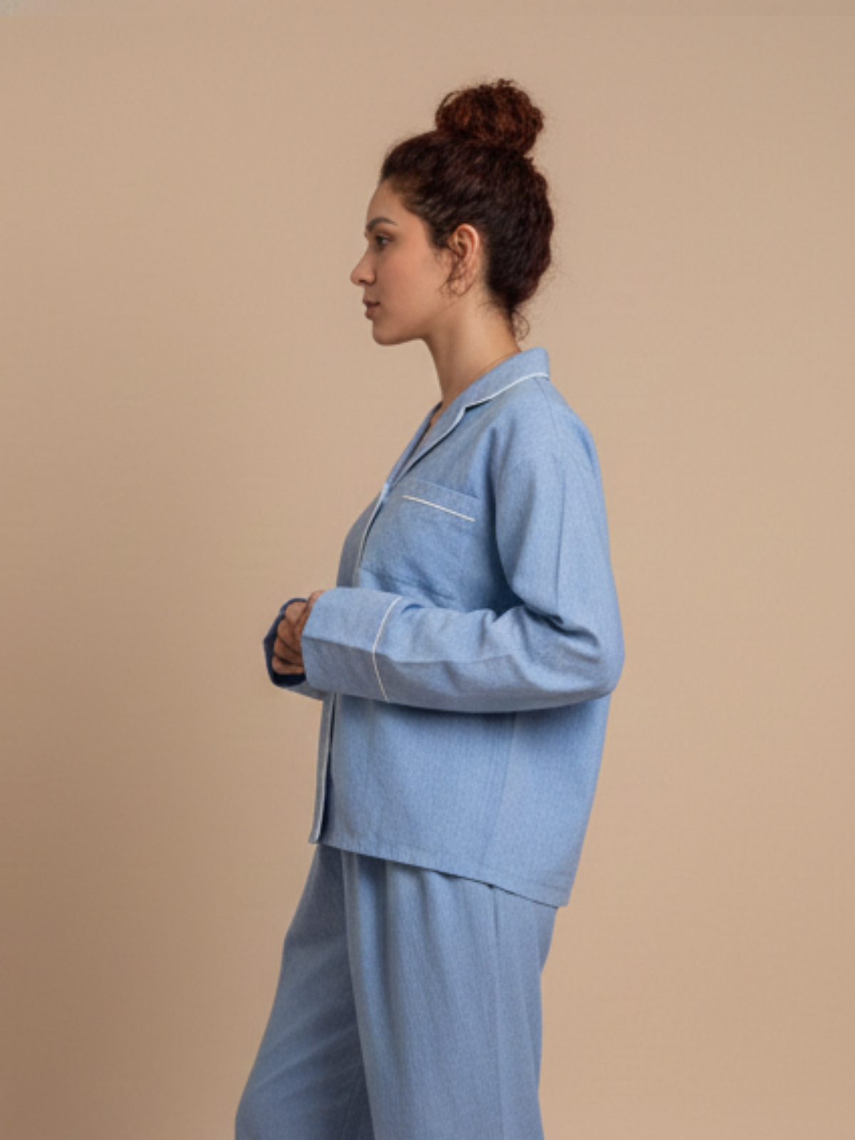 A woman in profile wearing a light blue pajama set with white piping details, standing against a solid beige background.