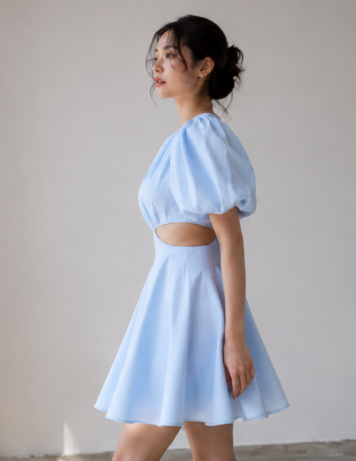 A woman wearing a light blue mini dress with a puff sleeve, a side cutout, and a flared skirt, standing against a plain light gray wall.