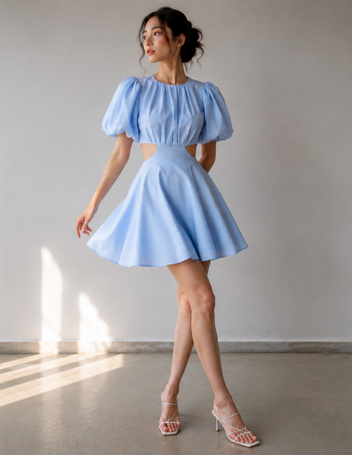 A woman modeling a light blue short dress with puff sleeves, a high-waisted cutout waist, and a flared skirt, paired with clear high-heeled sandals, standing in a minimalist studio setting.