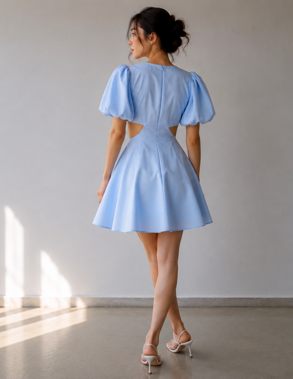A woman wearing a light blue mini dress with puff sleeves and side cutouts, shown from the back in a minimalist indoor setting with a light gray wall and concrete floor.