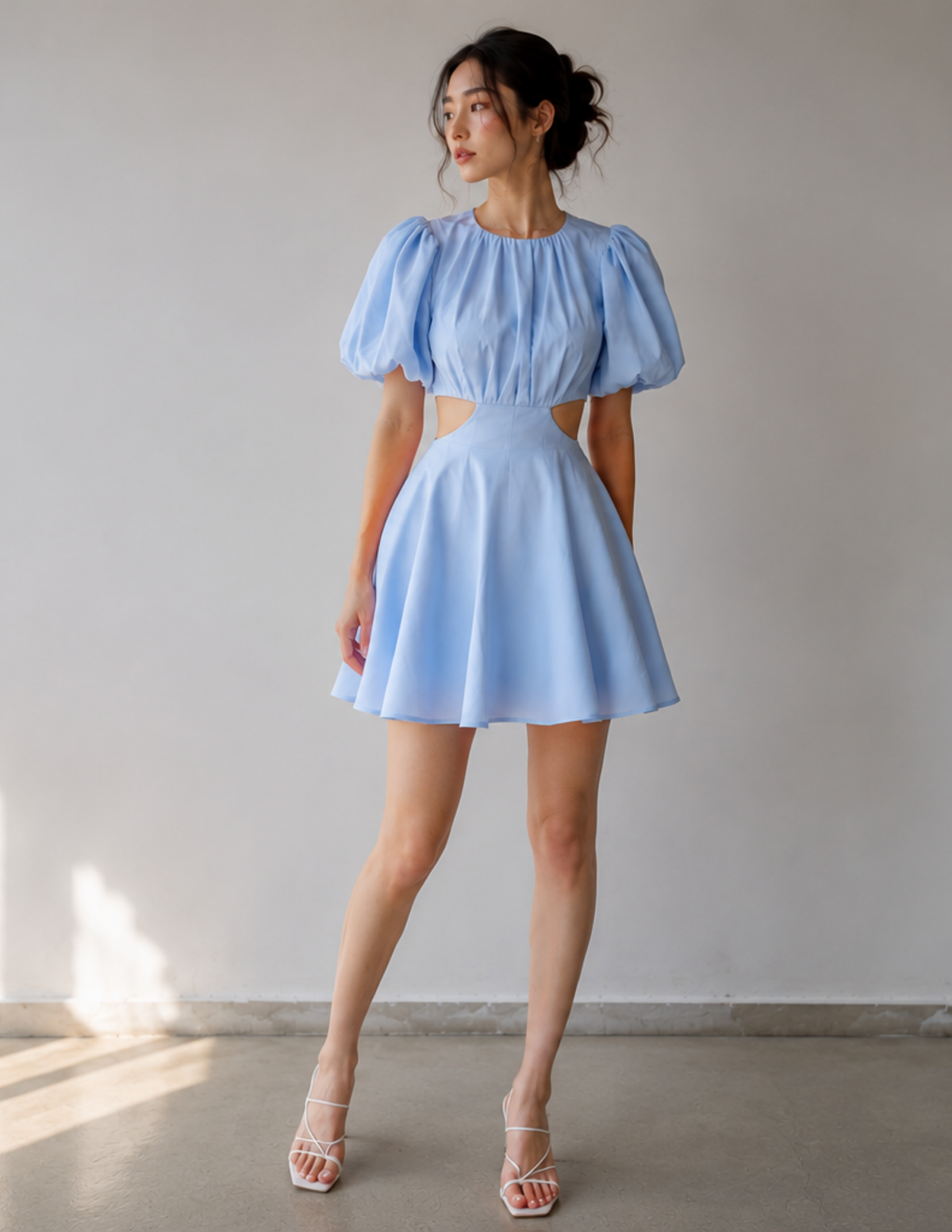 A woman modeling a light blue mini dress with puffed short sleeves, a high-waisted cut-out waist, and a flared skirt, paired with white strappy heels, set against a plain light gray wall.