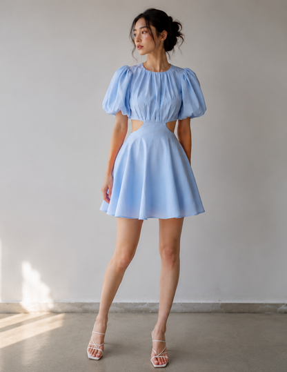 A woman modeling a light blue mini dress with puffed short sleeves, a high-waisted cut-out waist, and a flared skirt, paired with white strappy heels, set against a plain light gray wall.