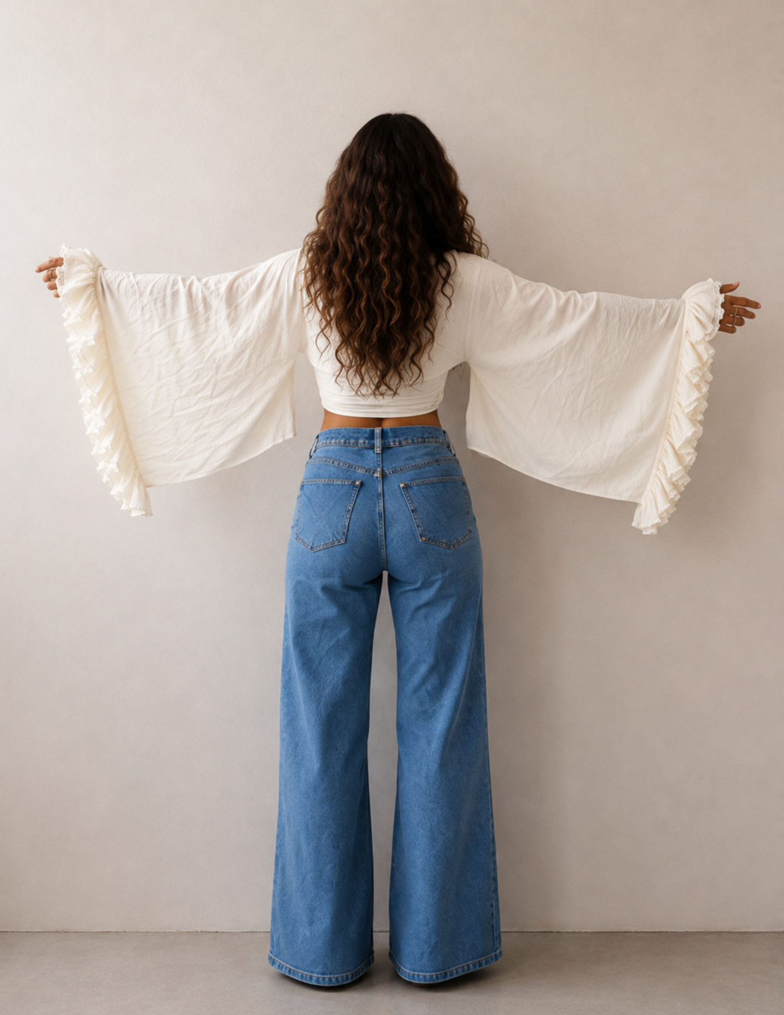 A woman viewed from behind wearing a cream-colored, long-sleeve bohemian-style top with ruffled edges and wide flared sleeves, paired with high-waisted blue wide-leg jeans, set against a plain beige wall.