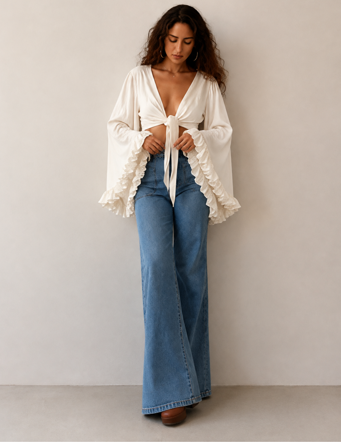 A woman stands against a plain light beige wall wearing a cream-colored, long-sleeve bohemian-style blouse with ruffled cuffs and a front tie, paired with high-waisted blue flare jeans.