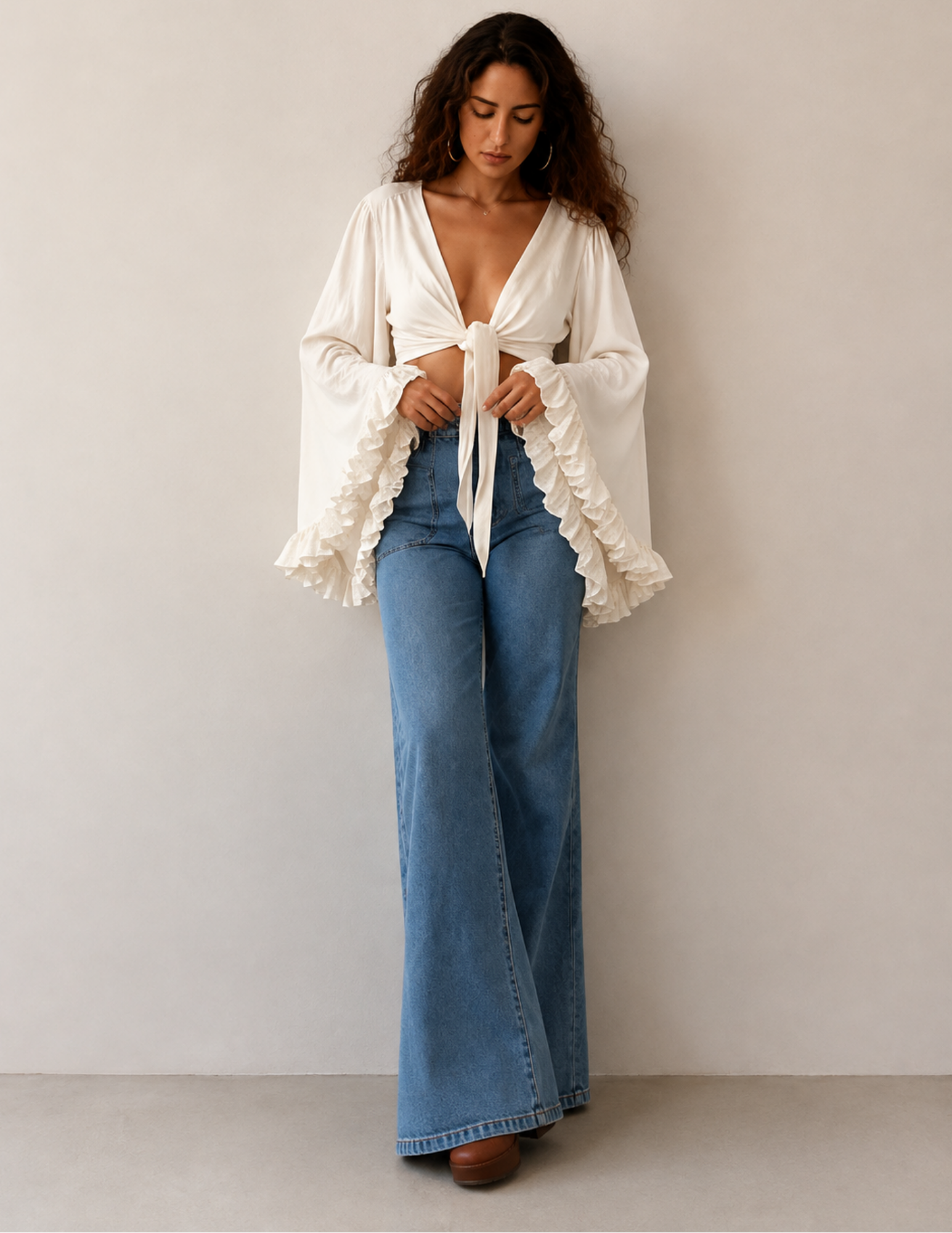 A woman stands against a plain light beige wall wearing a cream-colored, long-sleeve bohemian-style blouse with ruffled cuffs and a front tie, paired with high-waisted blue flare jeans.