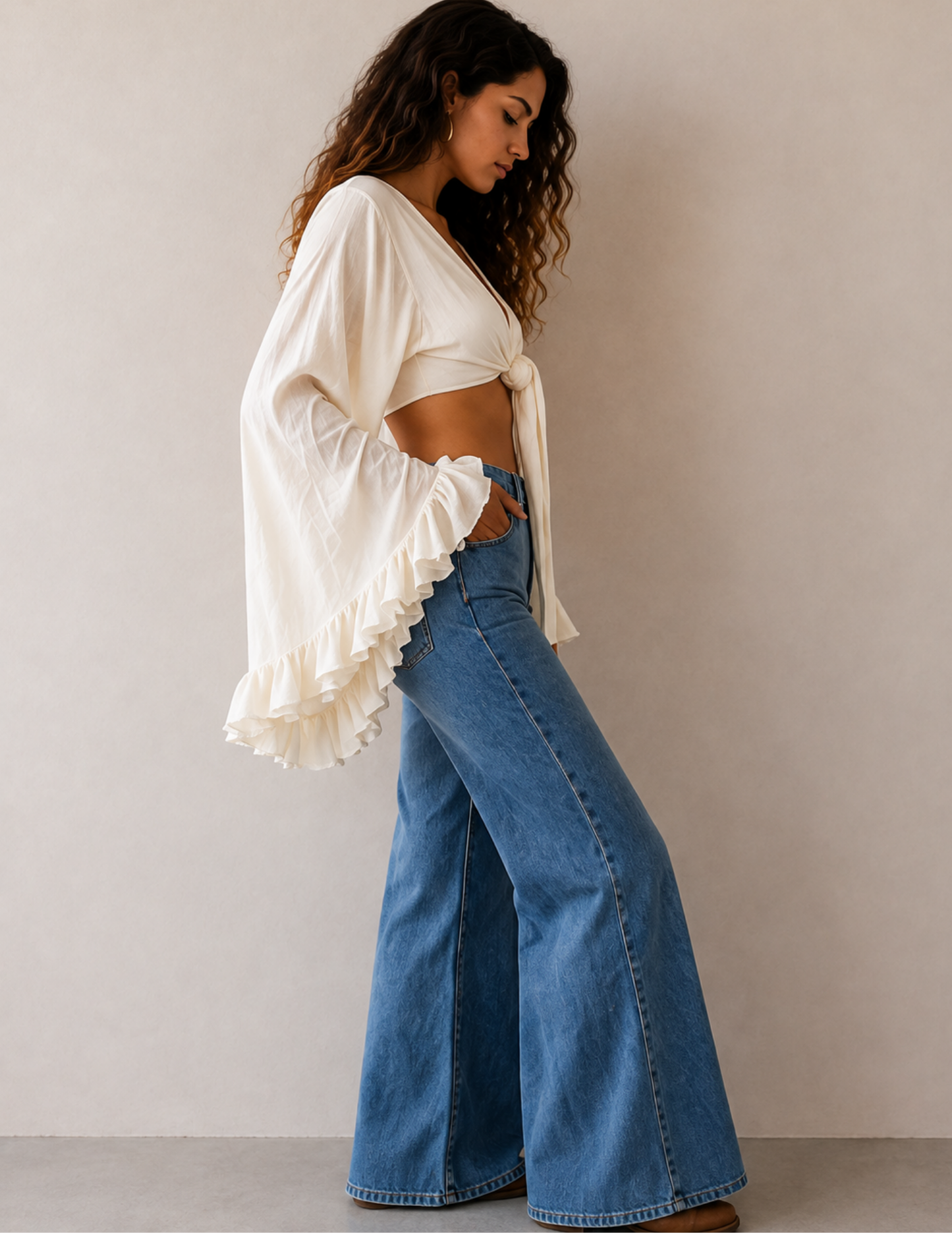 A woman wearing a cream ruffled long-sleeve crop top and wide-leg blue denim jeans, standing against a neutral beige wall.