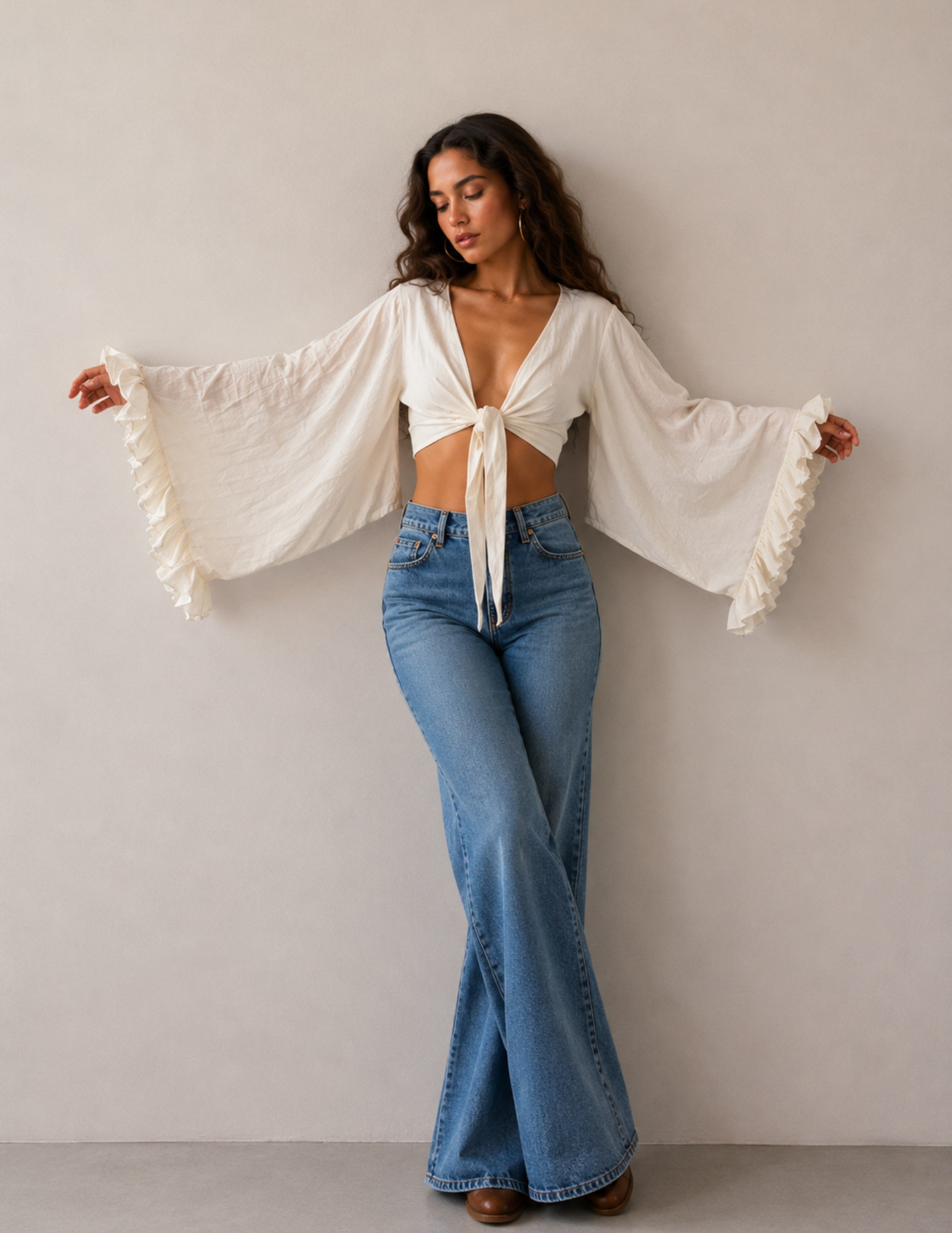 A woman modeling a white crop top with dramatic bell sleeves and ruffle trim, paired with blue flare jeans, standing against a plain light gray wall.