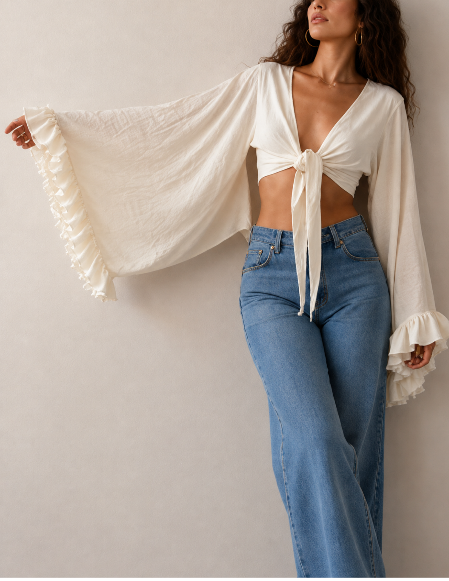 A woman models a cream-colored, long-sleeve tie-front crop top with dramatic ruffled bell sleeves, paired with high-waisted blue jeans, against a plain beige wall.