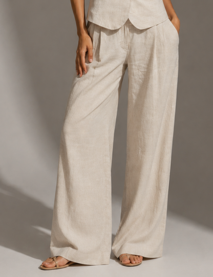 A person wearing loose, wide-leg linen pants in a light oatmeal beige color, paired with matching top and flat sandals, photographed against a neutral studio backdrop.