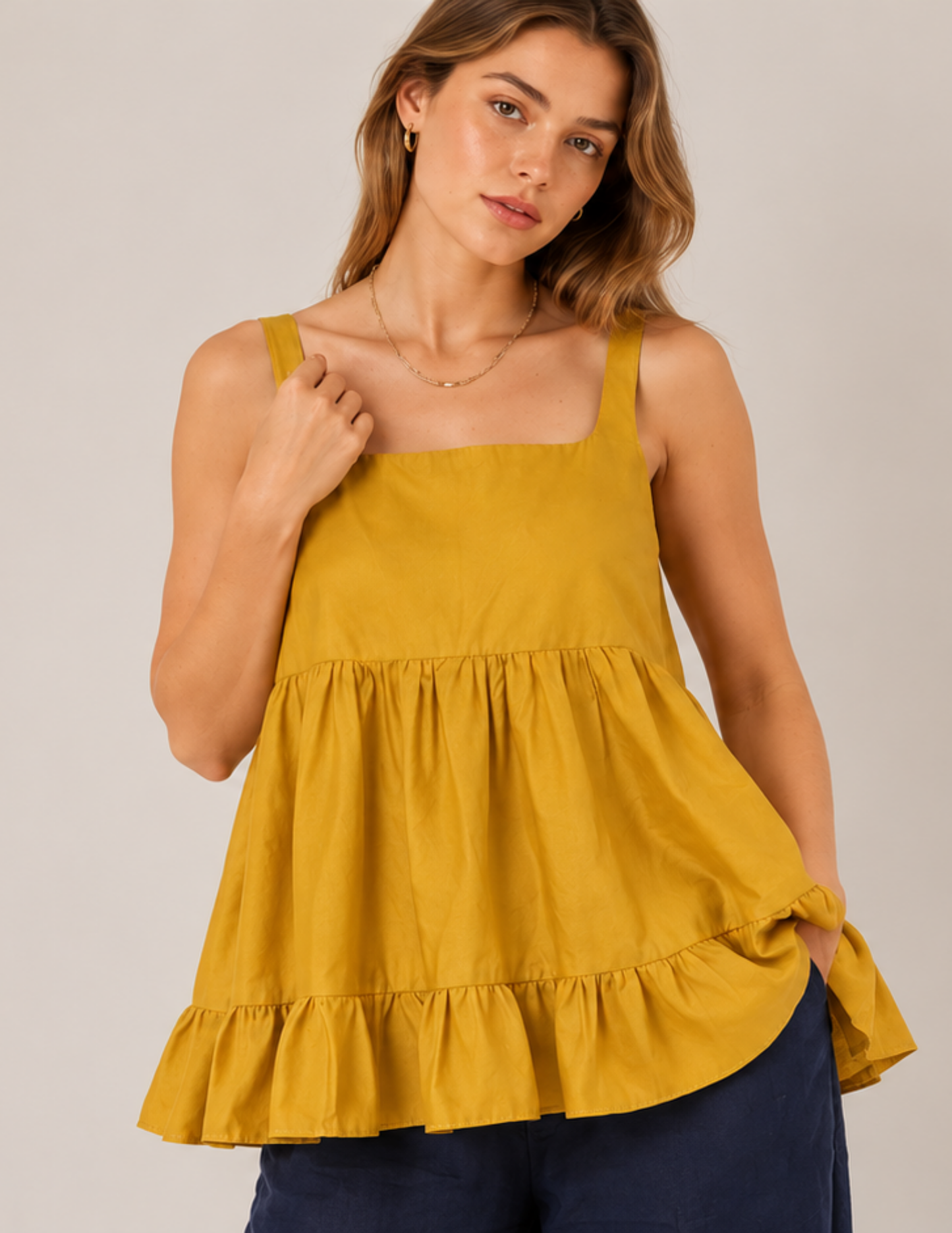A woman wearing a mustard yellow tiered peplum top with a square neckline and thin shoulder straps, paired with navy blue pants, against a solid light gray background.