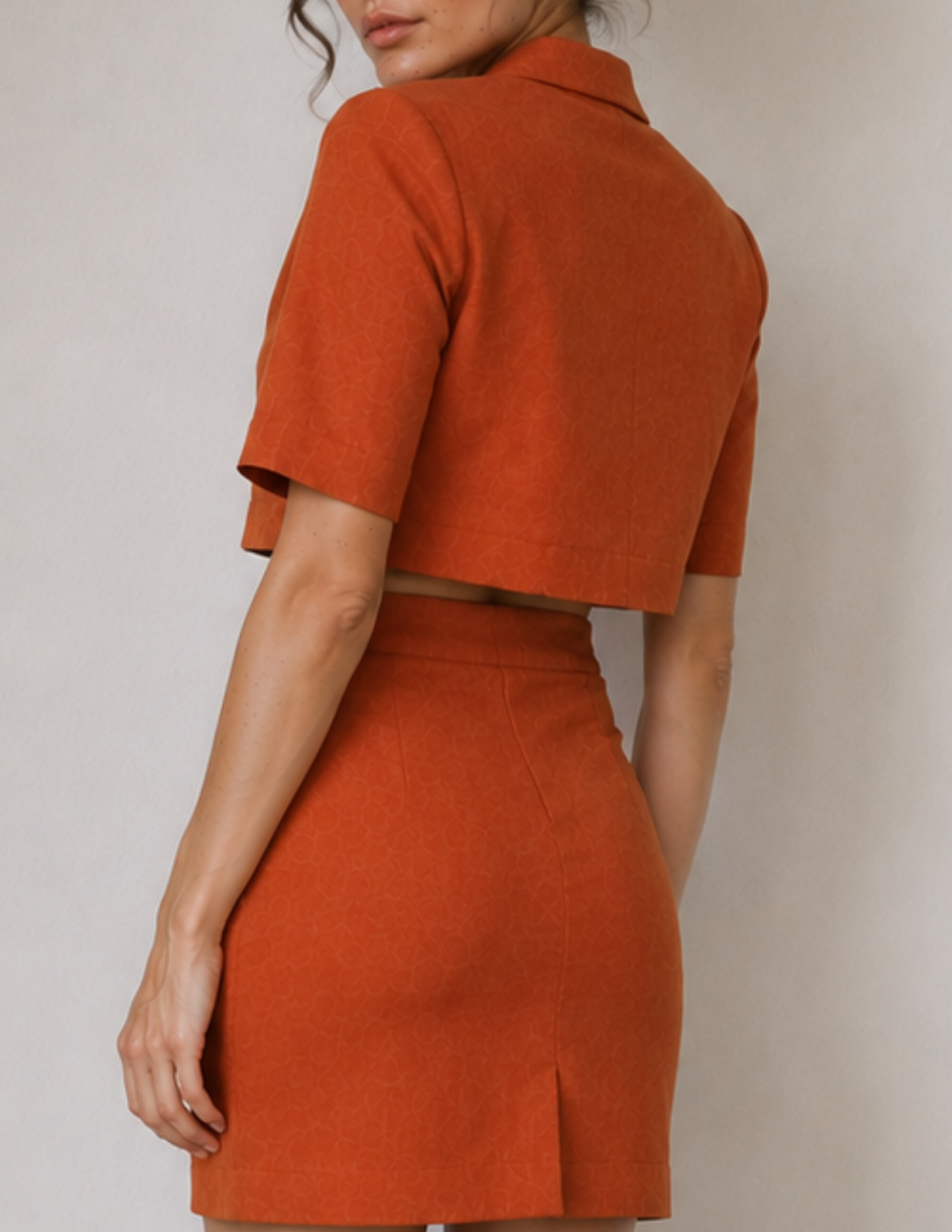 A woman wearing a matching burnt orange two-piece set consisting of a short-sleeve cropped collared top and a high-waisted mini skirt, shown from a rear three-quarter angle against a neutral light gray background.