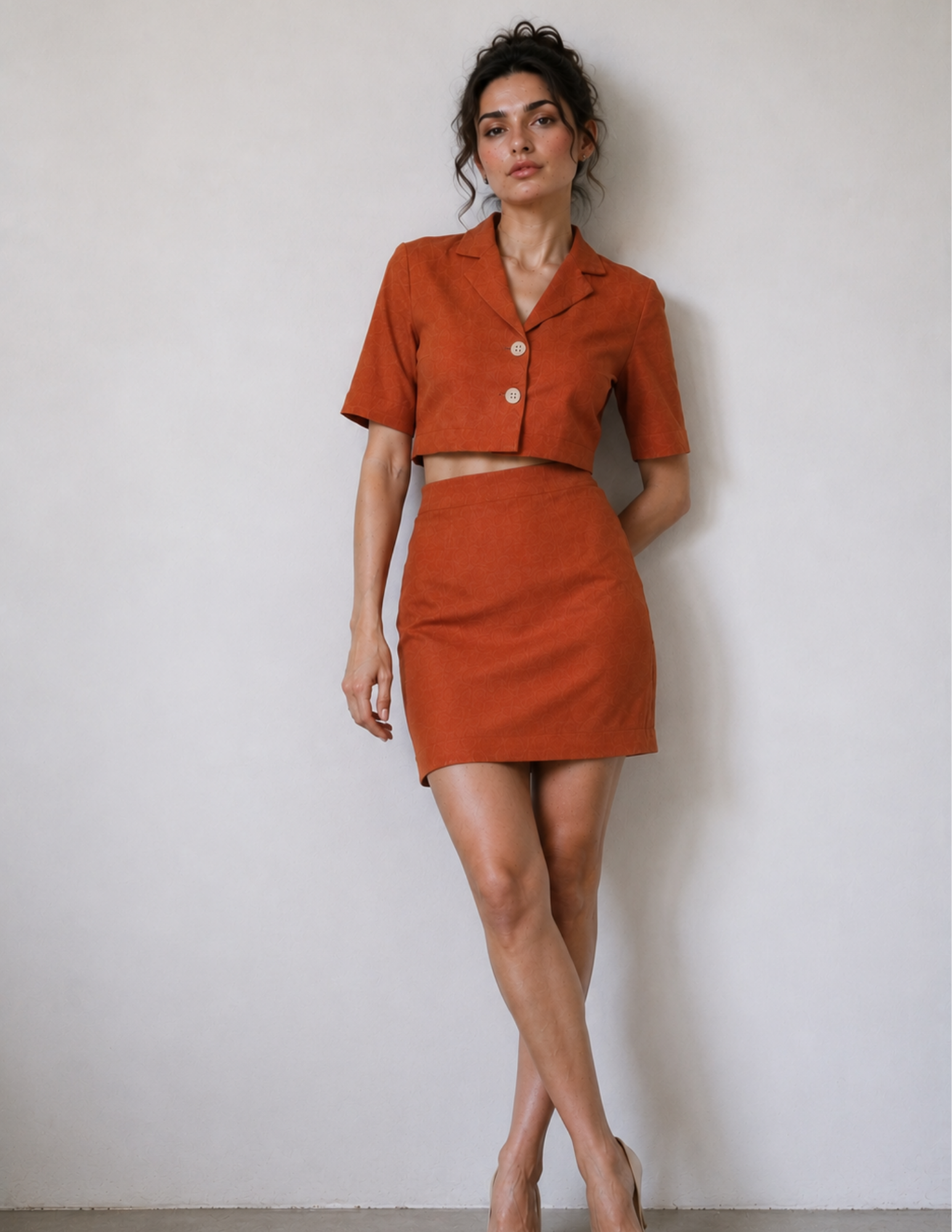 A woman modeling a matching burnt orange two-piece set consisting of a short-sleeve cropped button-front top and a high-waisted mini skirt, standing against a plain off-white wall.