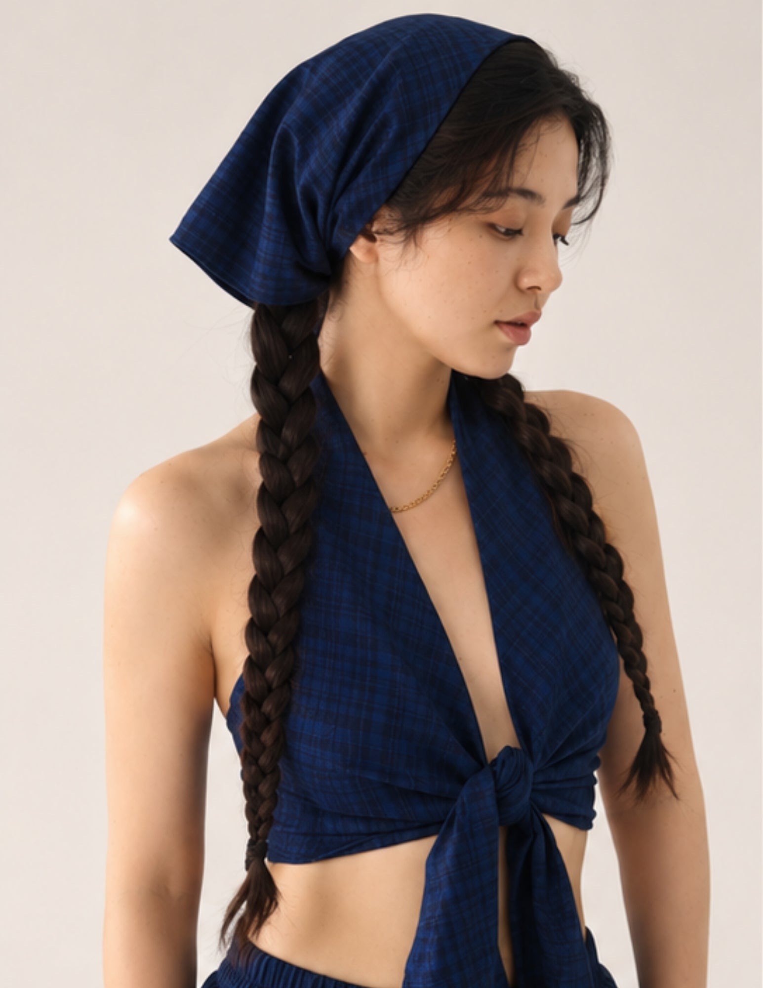 A woman wearing a dark blue plaid halter crop top and matching headscarf, with her hair styled in two long braids, photographed against a plain light beige studio background.