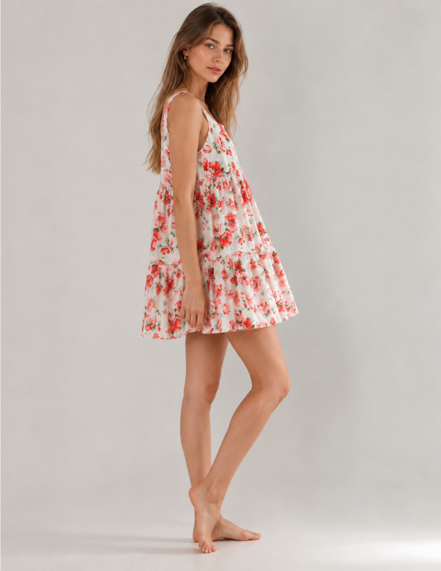 A woman modeling a sleeveless tiered mini dress with a red and green floral print on a white background, photographed against a neutral light gray studio backdrop.