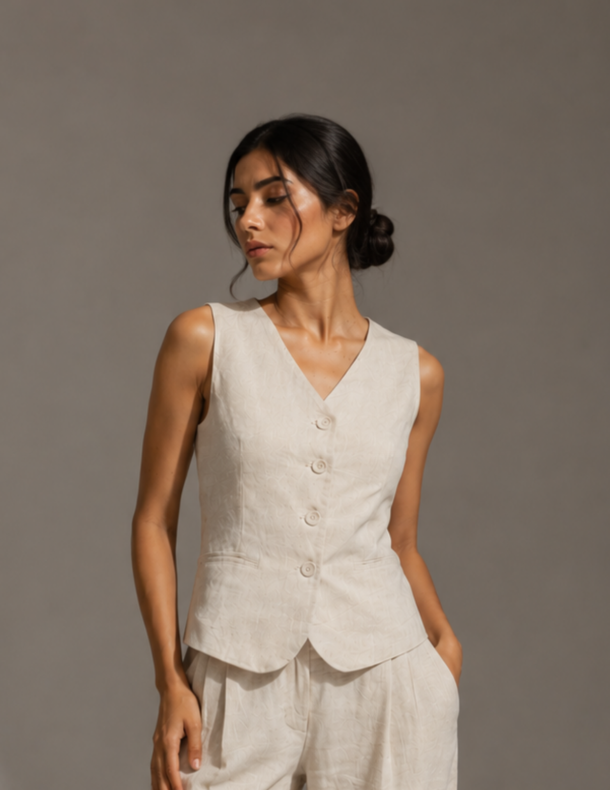 A woman modeling a sleeveless light beige linen vest with a button front and matching trousers, set against a solid gray studio background.