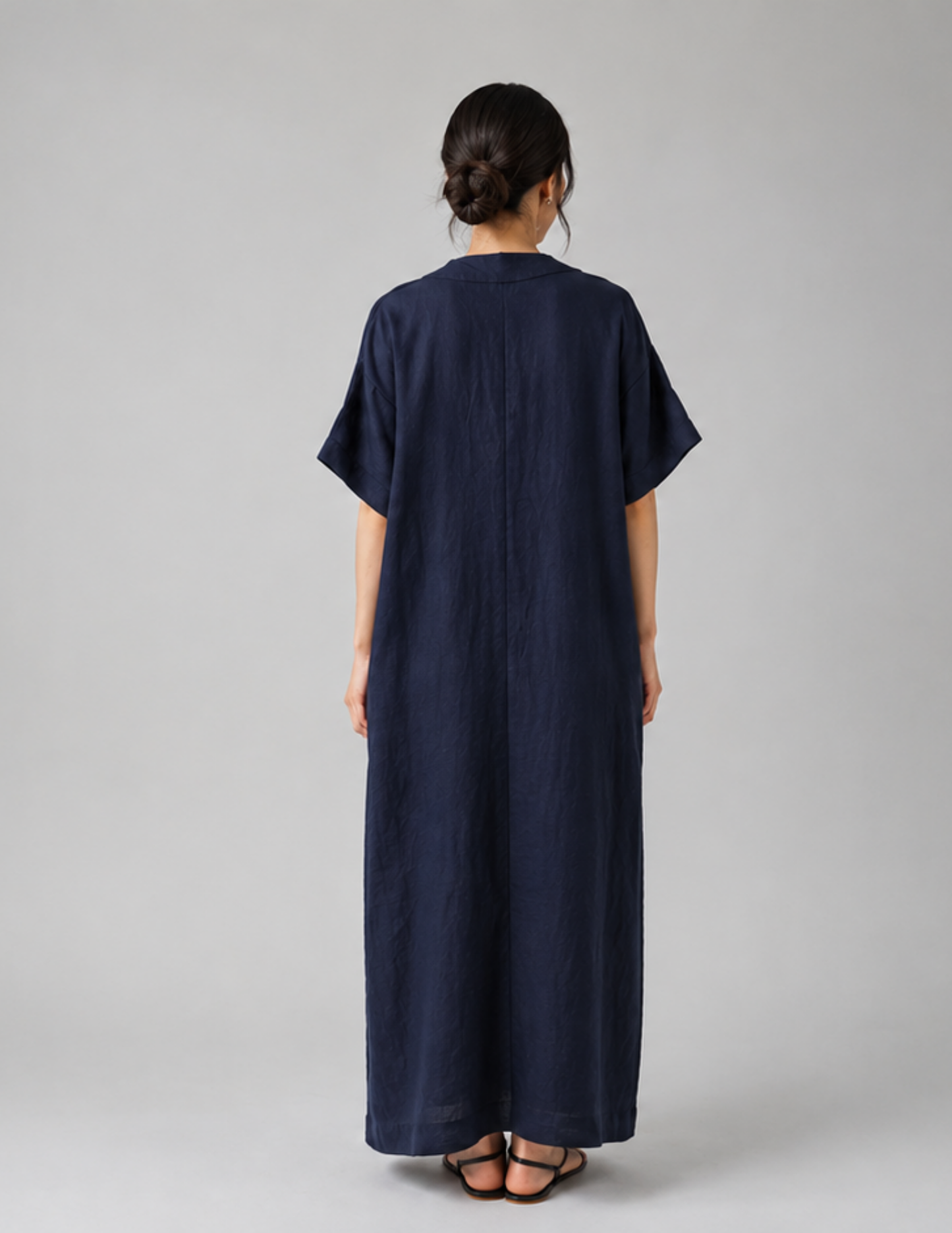 A woman viewed from the back wearing a loose, dark navy maxi dress with short sleeves and sandals, standing against a plain light gray studio background.