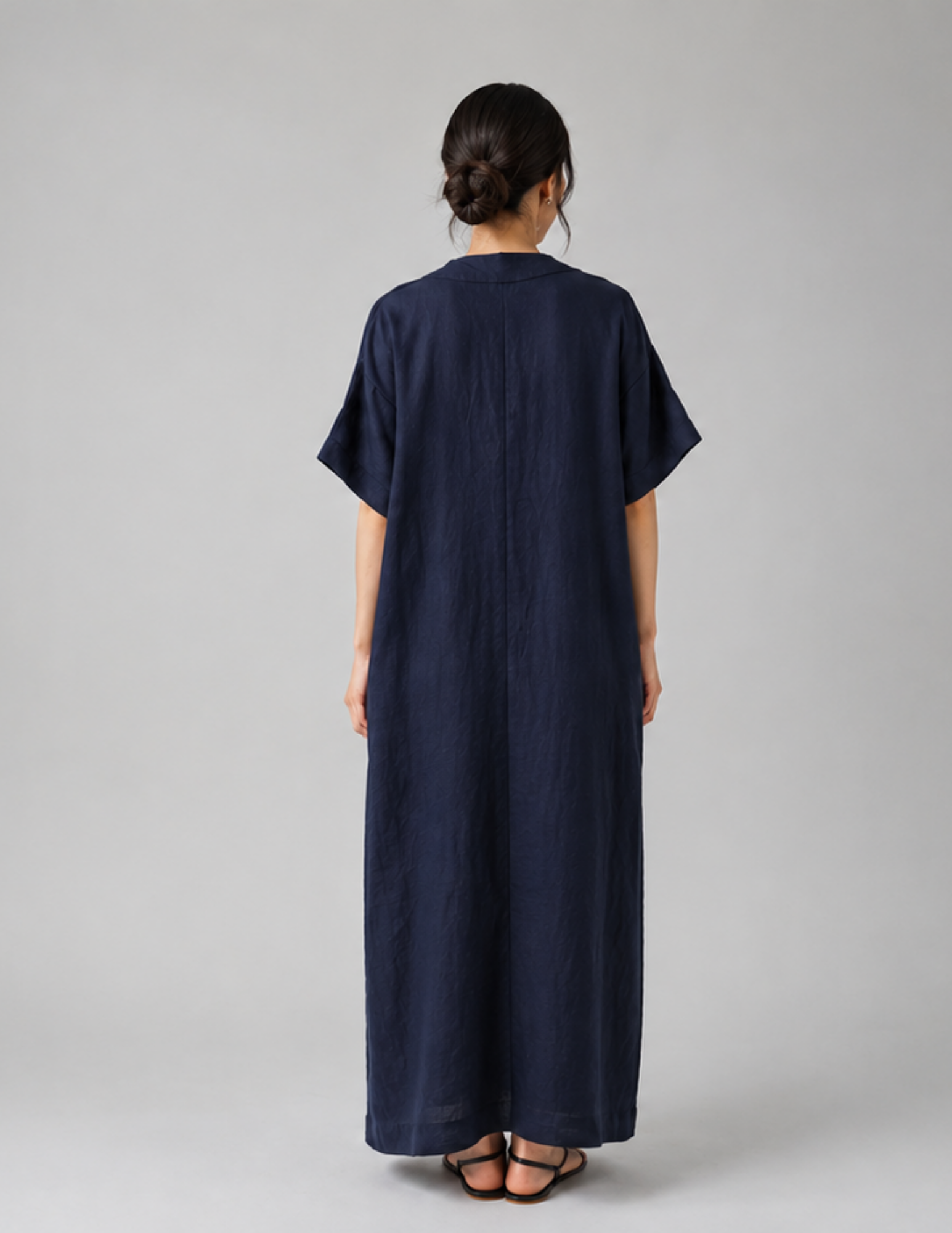 A woman viewed from the back wearing a loose, dark navy maxi dress with short sleeves and sandals, standing against a plain light gray studio background.