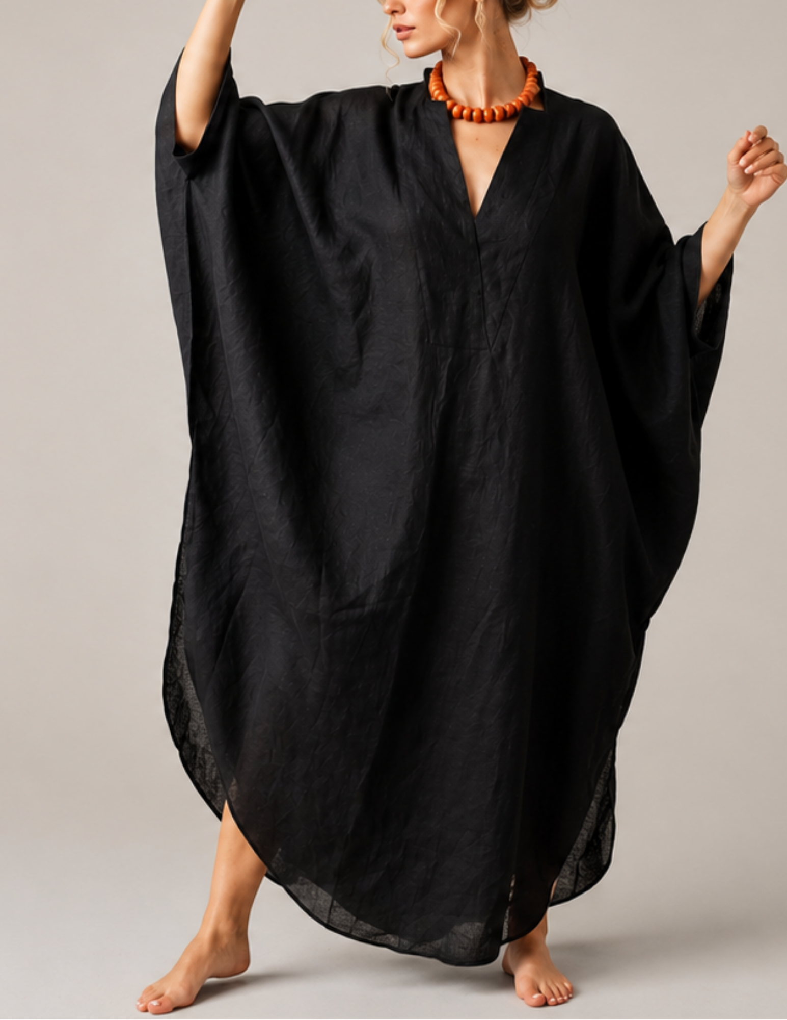 A woman modeling a loose, black, kaftan-style dress with wide draped sleeves and a deep V-neckline, set against a neutral light gray studio background.